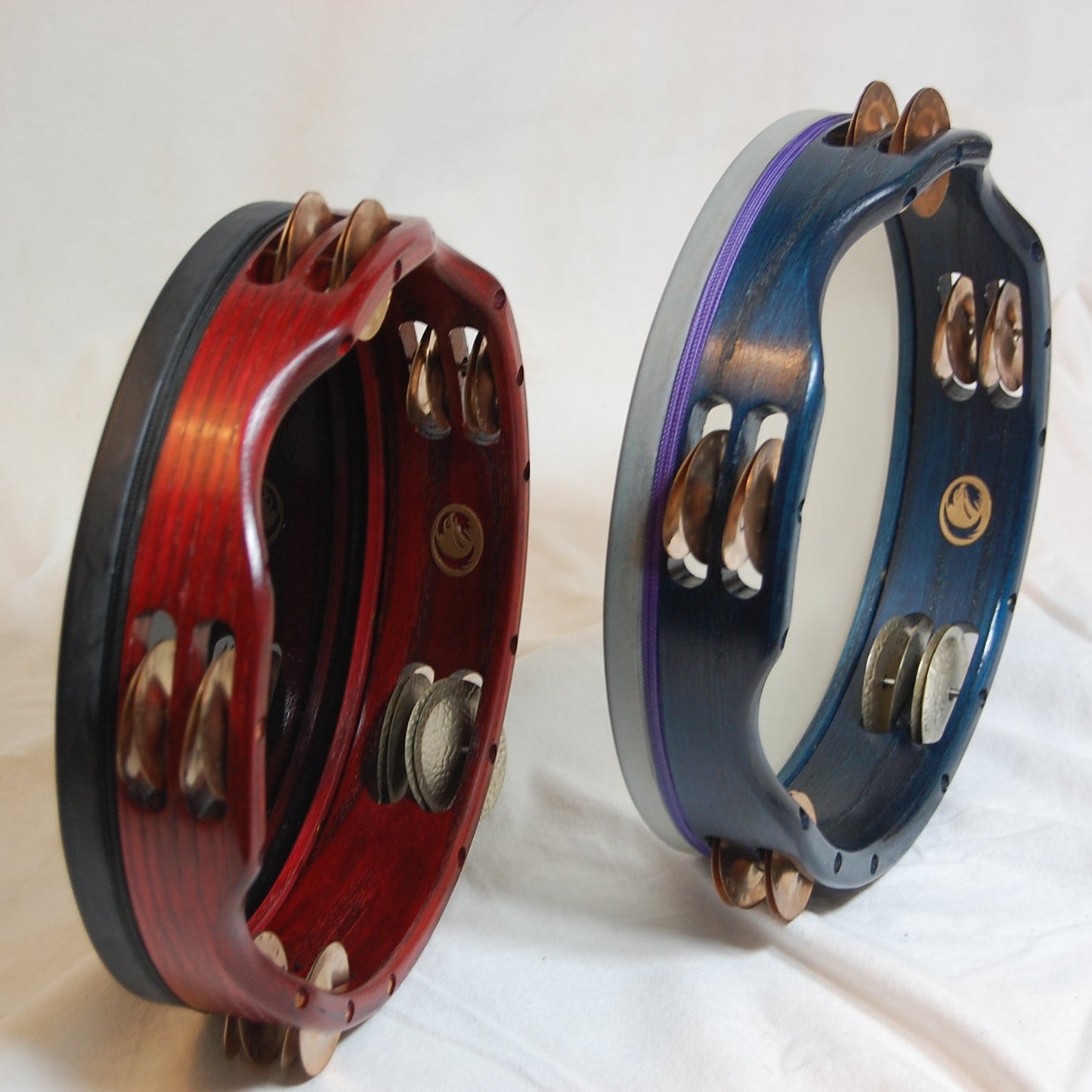 Two Inanna Tambourines with visible copper and nickel jingles, one with a red stain and the other with a blue stain, both with a clear lacquer overcoat.