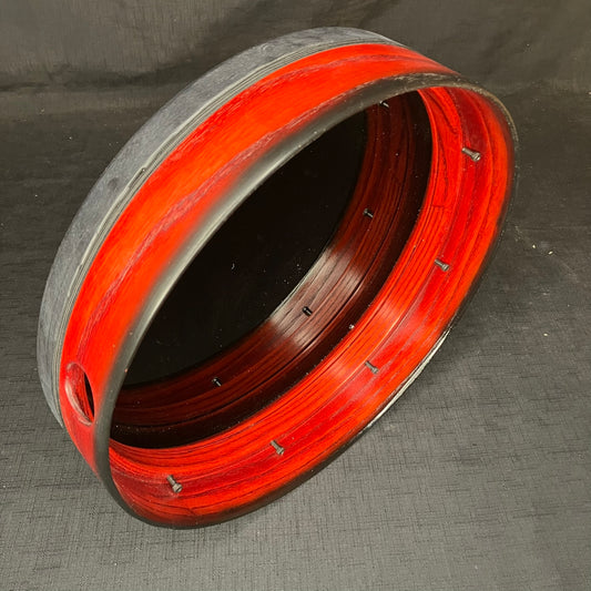 A 14-inch diameter frame drum with a cherry red stained finish and black fade edge, featuring a thumbhole grip and black cording, without a drum head.