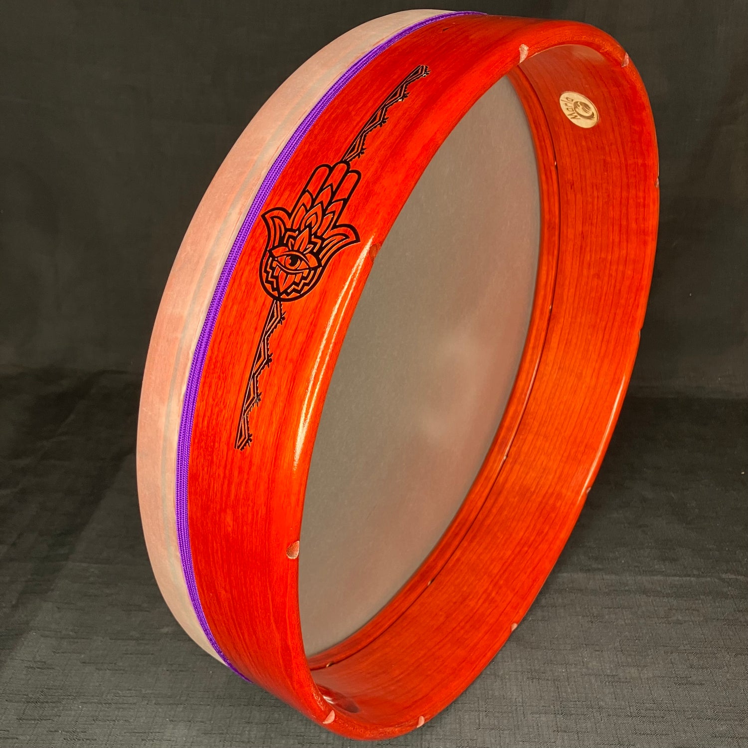 A 16-inch Marla Leigh Series Tar Frame Drum with a red and orange stained shell, a thumb hole, and a decorative graphic of a hand.