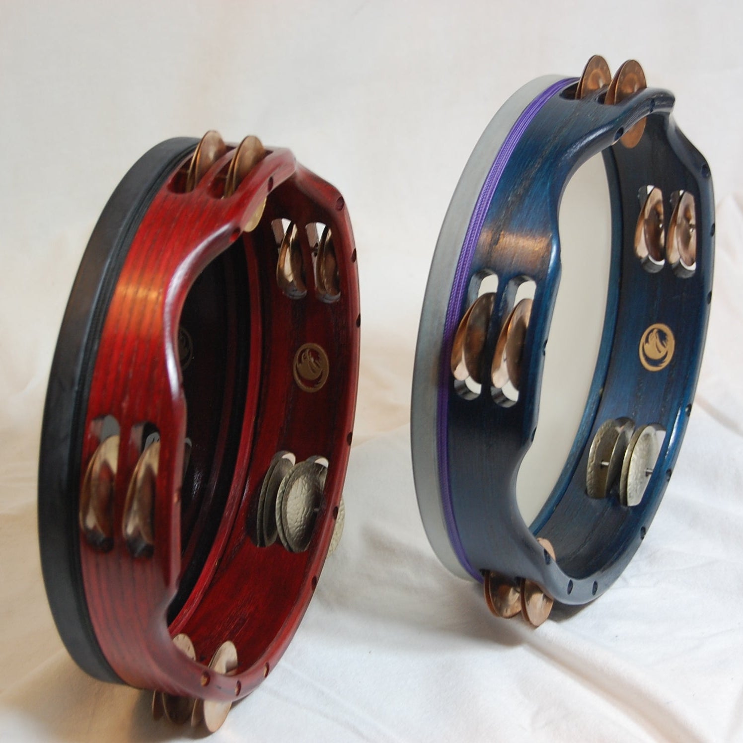 Two Inanna Tambourines with visible copper and nickel jingles, one with a red stain and the other with a blue stain, both with a clear lacquer overcoat.