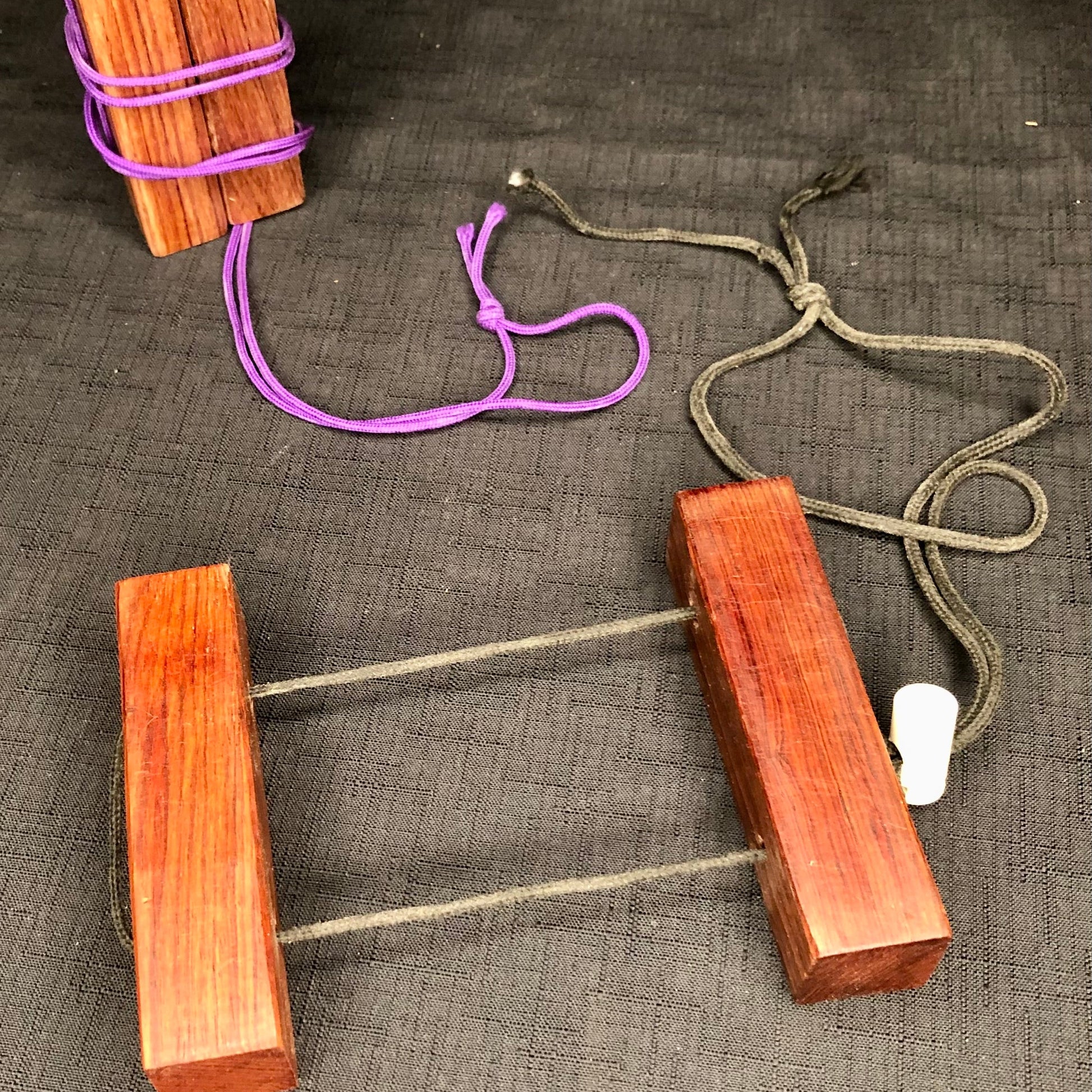 An adjustable roll-stop device made of rosewood, comprising two hardwood blocks connected by a purple adjustable lanyard, designed to stabilize a drum.