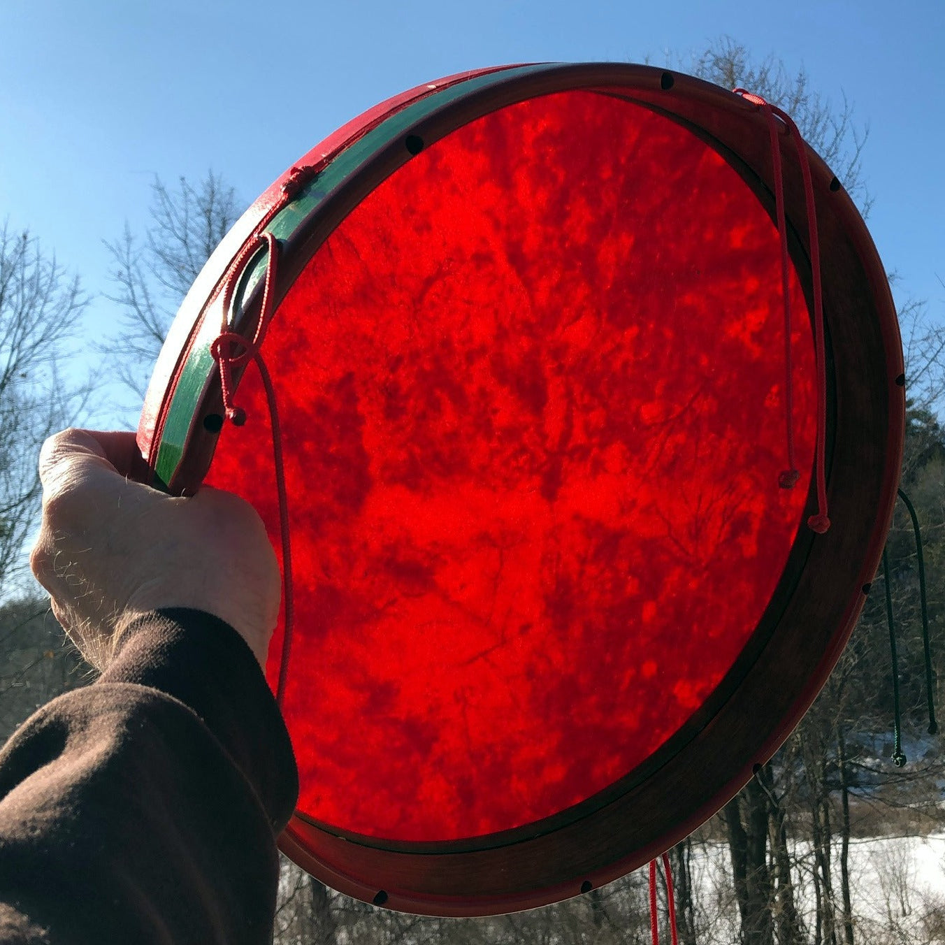 A person holding a tympanum drum with a goatskin head and green wood body, featuring five knotted cords.